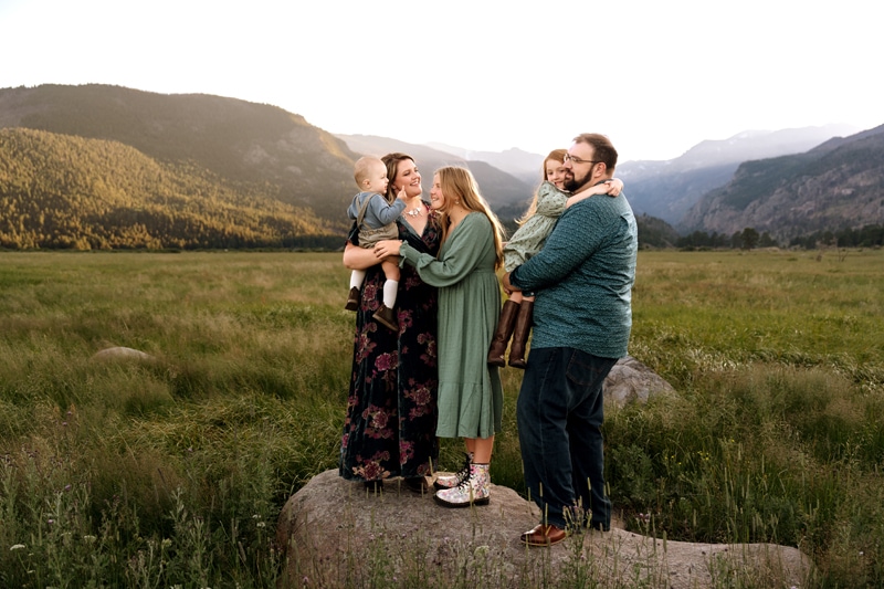 Family Photographer, a family of 5 huddles close before meadows and mountains, mom holds baby