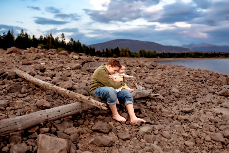 Family Photographer, a boy sits on a log near the lake with baby sibling