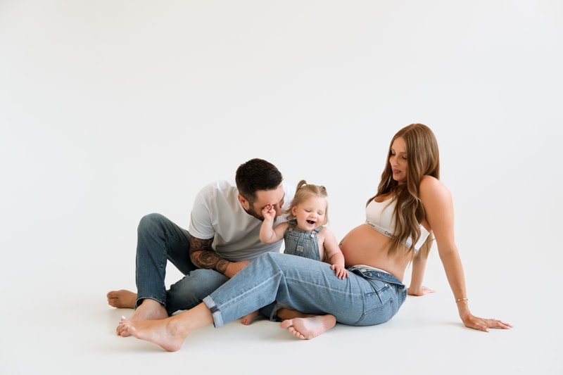 Family Photographer, a pregnant woman sits near her husband and baby girl