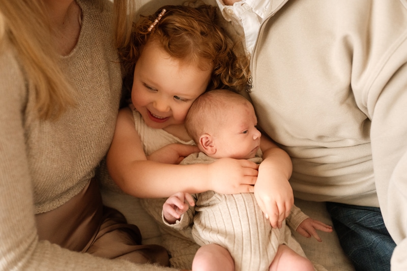 Family Photographer, a little girl holds her new baby sibling and smiles in some cushions