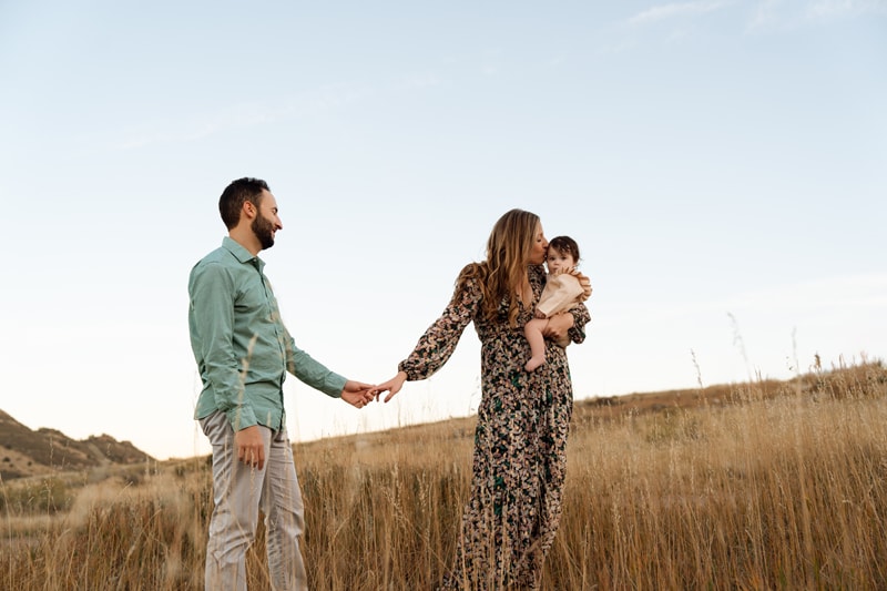 Family Photographer, a husband and wife hold hands in a dry meadow, she kisses her baby in her arms